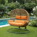 Balançoire Orvella 2 places avec coussin orange dans un jardin, cadre métal noir et panier en rotin synthétique